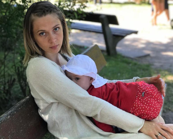 Eine Frau sitzt auf einer Parkbank und stillt ihr Kleinkind, schaut etwas genervt in die Kamera.