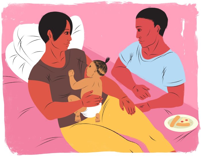 Eine Mutter stillt ihr Baby auf einem Bett, ihr Mann sitzt daneben, sie schauen sich wohlwollen an.